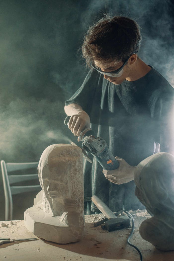 An artist skillfully carves a stone sculpture using power tools amidst a haze of dust.